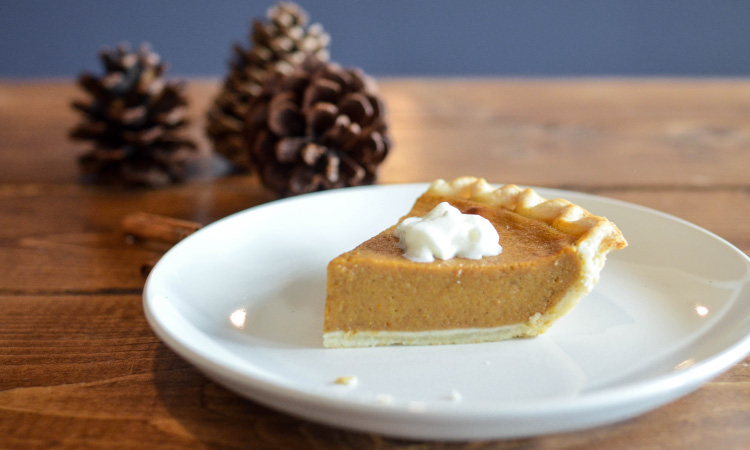 Closeup of a piece of pumpkin pie with a dollop of whipped cream on a white plate next to a pie of pine cones