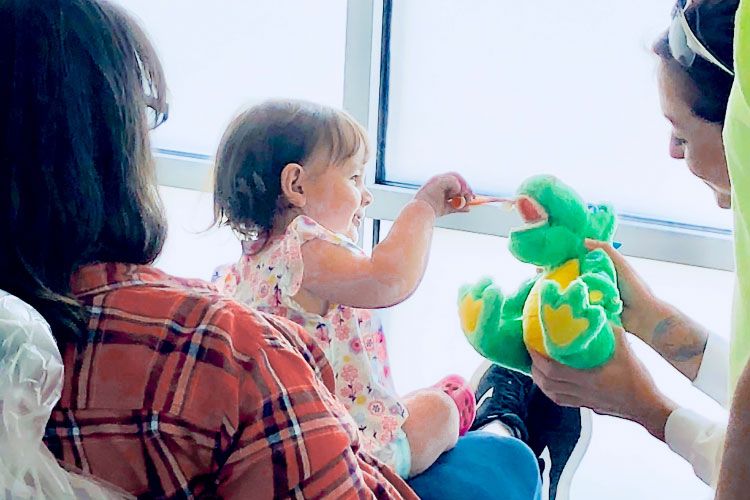Young girl sits on her mother's lap at the dentist's office brushing the teeth of a stuffed dinosaur