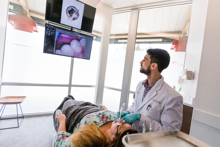 Dr. Perlman examines the teeth of a blonde female patient with an image of her teeth on a TV screen