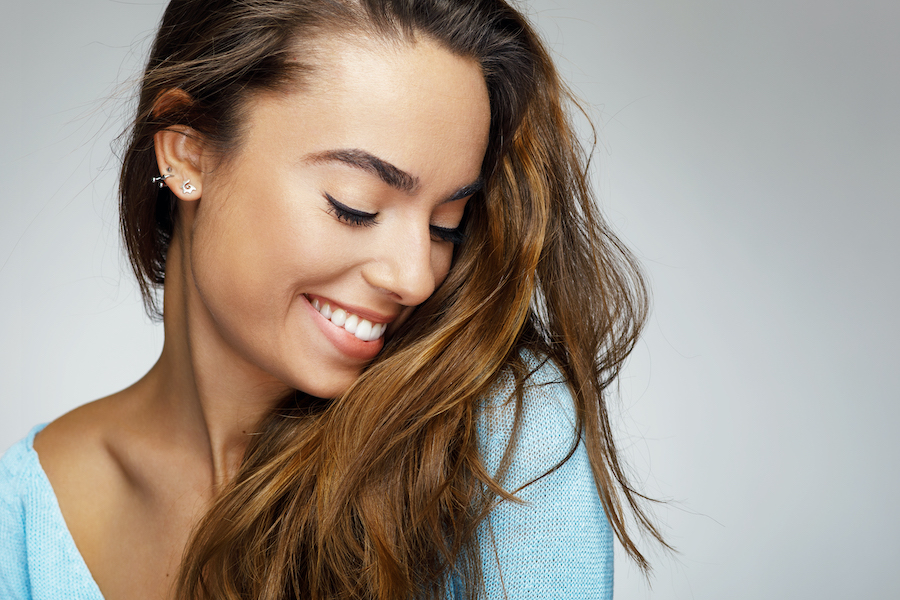 Brunette woman smiles and looks down after getting dermal fillers to plump and smooth her skin