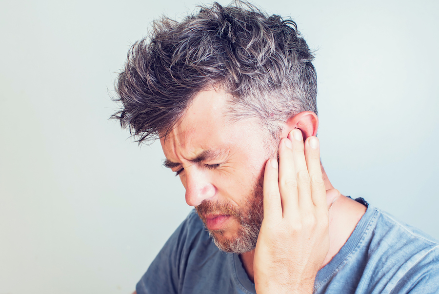 Middle-aged man in a gray shirt cringes in discomfort and touches his ear due to jaw pain